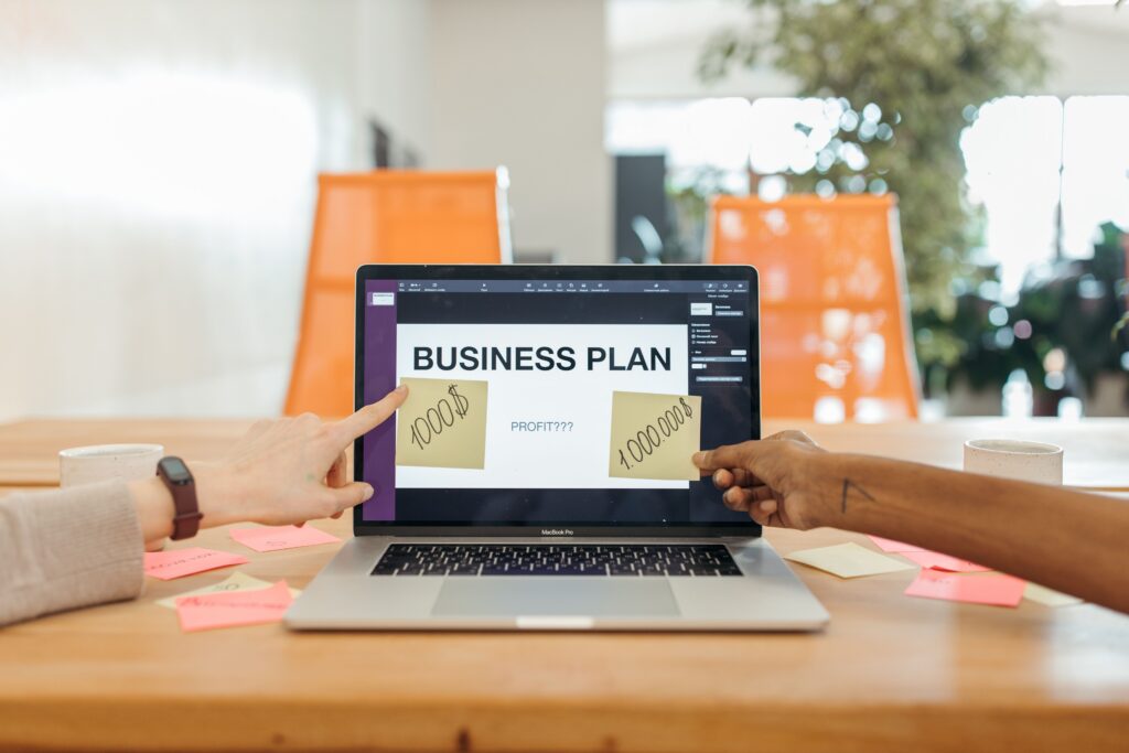 A laptop with the words Business Plan showing on screen and some post it notes with numbers and profit written on it